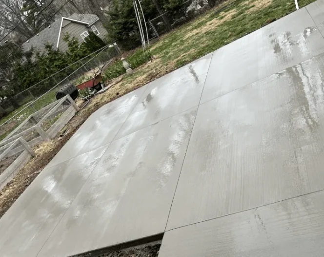 A newly poured concrete patio, divided into large squares, with visible brush strokes, next to a grassy backyard.